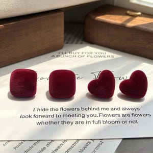 May include: Four small, deep red velvet-textured objects arranged in a row on a white card with printed text. Two objects are square-shaped with rounded corners, and two are heart-shaped. The text reads: I'll buy for you a bunch of flowers.