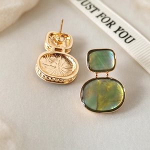 May include: A pair of gold-tone stud earrings featuring a rectangular top and an oval bottom, both inlaid with iridescent green shell-like material. One earring shows the back post, while the other displays the front surface. A ribbon reads JUST FOR YOU.