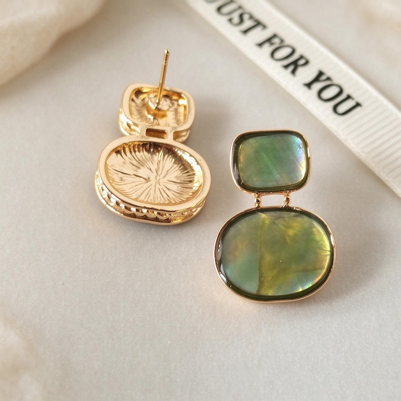 May include: A pair of gold-tone stud earrings featuring a rectangular top and an oval bottom, both inlaid with iridescent green shell-like material. One earring shows the back post, while the other displays the front surface. A ribbon reads JUST FOR YOU.