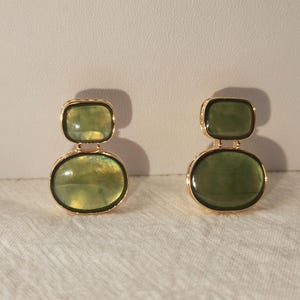 May include: A pair of drop earrings featuring two stacked, smooth, olive-green translucent stones set in gold-toned metal frames. The top stone is rectangular with rounded corners, while the bottom stone is oval-shaped.