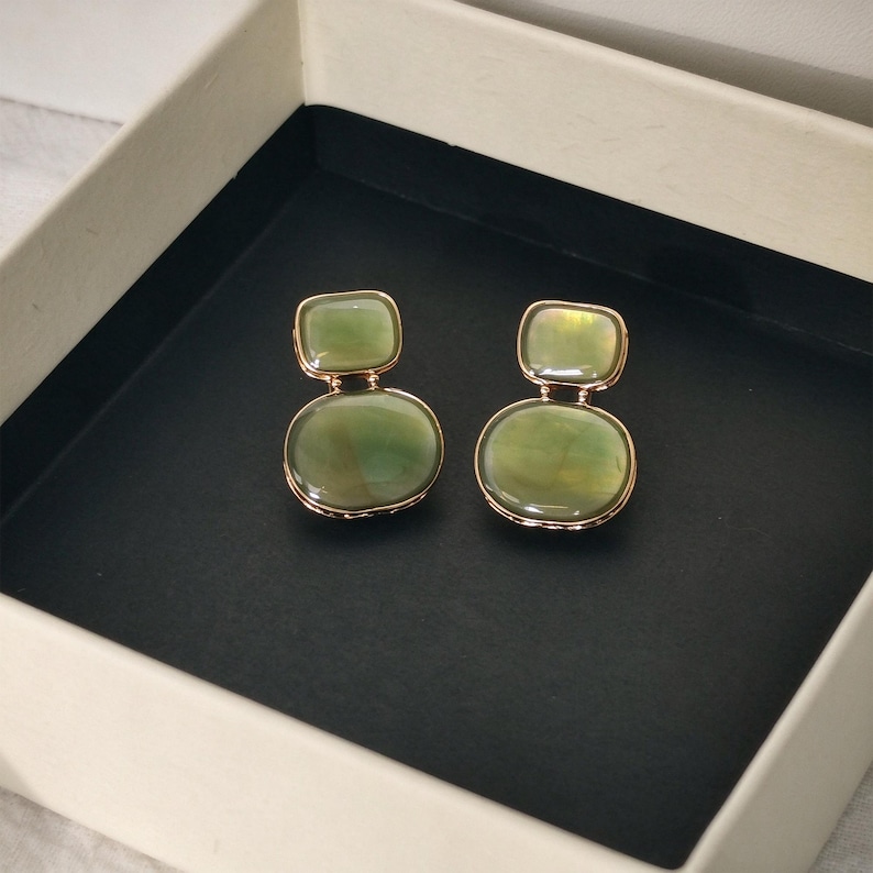 May include: A pair of drop earrings featuring two smooth, polished green stones set in gold-toned metal. Each earring consists of a smaller rectangular stone positioned above a larger oval stone, connected by a thin gold-toned metal link.