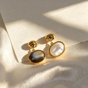 May include: A pair of gold-toned earrings featuring a textured, hammered-finish circular top piece connected to an oval-shaped pendant. One pendant contains a dark, iridescent gray stone, while the other contains a white, iridescent stone.