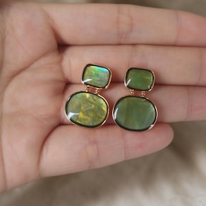 May include: A pair of drop earrings featuring two stacked, smooth, olive-green stones set in gold-toned metal. The top stone is a smaller rectangle with rounded corners, while the bottom stone is a larger, rounded oval shape.