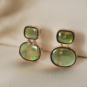 May include: A pair of gold-toned drop earrings featuring two stacked, iridescent green stones. The top stone is rectangular with rounded corners, while the bottom stone is oval. Each stone is encased in a thin black border and gold metal.