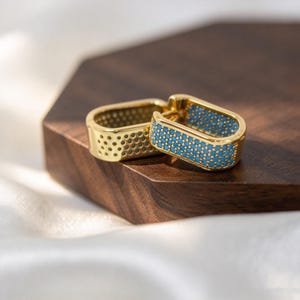 May include: A pair of gold-toned hoop earrings with a rectangular shape. The outer surface of each hoop is inlaid with small, bright turquoise blue stones arranged in a grid pattern, creating a textured appearance against the polished gold metal.