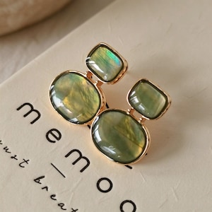May include: A pair of gold-toned earrings featuring two stacked, smooth, oval-shaped green iridescent stones. The earrings are resting on a light-colored surface with the partial black text "memo" printed on it.