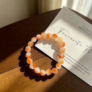 Könnte beinhalten: Ein Armband aus facettierten, durchscheinenden orangefarbenen und weißen Perlen. Die Perlen haben eine geometrische, mehrseitige Form und sind kreisförmig aufgereiht. Das Armband liegt neben einer weißen Karte mit schwarzem Textaufdruck.