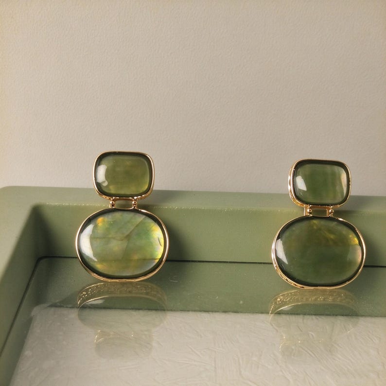 May include: A pair of gold-toned drop earrings featuring two stacked, smooth, translucent olive-green stones. The top stone is rectangular with rounded corners, while the bottom stone is larger and oval-shaped, both set in thin gold metal frames.