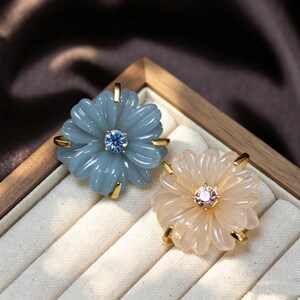 May include: Two flower-shaped brooches with gold-tone metal prongs. One brooch features translucent blue petals with a central blue rhinestone, while the other has translucent pale peach petals with a central pink rhinestone.