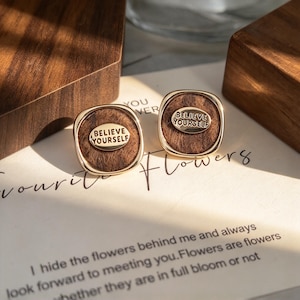 May include: A pair of gold-toned square stud earrings featuring a brown textured center inlay. Each earring has a small oval gold plaque in the middle with the embossed text "BELIEVE YOURSELF" in capital letters.