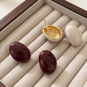May include: A set of four stud earrings with smooth, oval-shaped fronts. Two earrings are deep burgundy, one is cream-colored, and one is white with a gold-toned interior featuring a ribbed texture and a metal post backing.