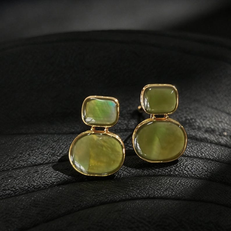 May include: A pair of gold-toned earrings featuring two stacked, smooth, olive-green stones. The top stone is rectangular with rounded corners, while the bottom stone is oval. Both stones exhibit a subtle, iridescent sheen within the setting.