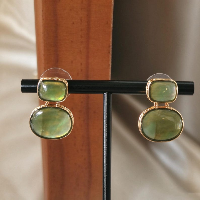 May include: A pair of gold-toned drop earrings featuring two stacked, smooth, translucent olive-green stones. The top stone is rectangular with rounded corners, while the bottom stone is oval. Each piece is set in a polished gold metal frame.