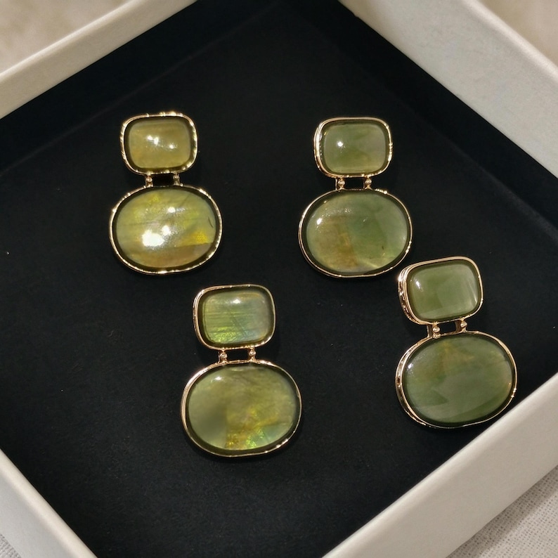 May include: Two pairs of drop earrings featuring smooth, polished olive-green stones set in gold-toned metal. Each earring consists of a smaller rectangular stone positioned above a larger oval stone, creating a balanced, layered appearance.