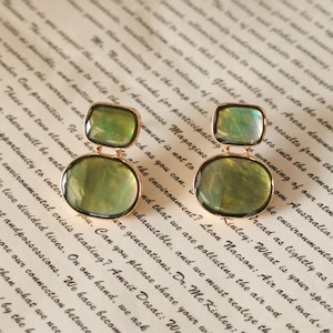 May include: A pair of gold-toned drop earrings featuring two stacked, smooth, translucent green stones. The top stone is a smaller square shape, while the bottom stone is a larger oval shape, both encased in a thin gold metal frame.