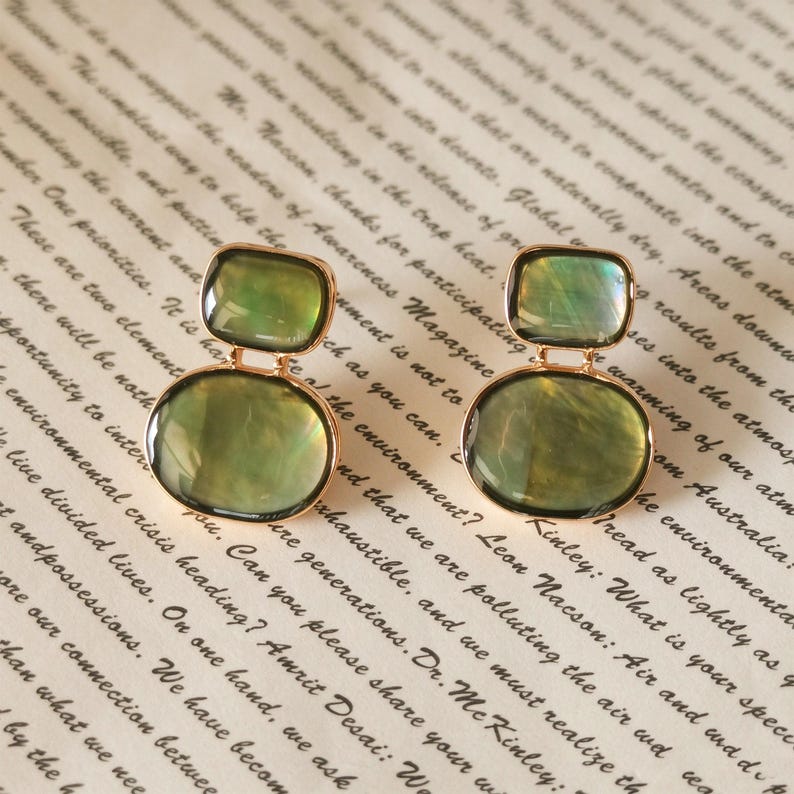 May include: A pair of gold-toned drop earrings featuring two stacked, smooth, translucent green stones. The top stone is a smaller square shape, while the bottom stone is a larger oval shape, both encased in a thin gold metal frame.