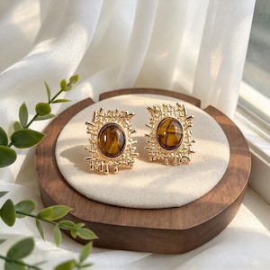 May include: A pair of gold-toned earrings featuring a rectangular, textured, open-work metal frame surrounding a central, smooth, oval-shaped brown and gold tiger's eye gemstone. The earrings have a polished finish and a detailed, irregular lattice design.