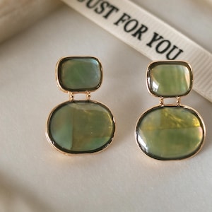 May include: A pair of gold-toned earrings featuring two stacked, smooth, olive-green iridescent stone elements. The top piece is rectangular with rounded corners, while the bottom piece is oval. The metal settings are thin and polished gold.