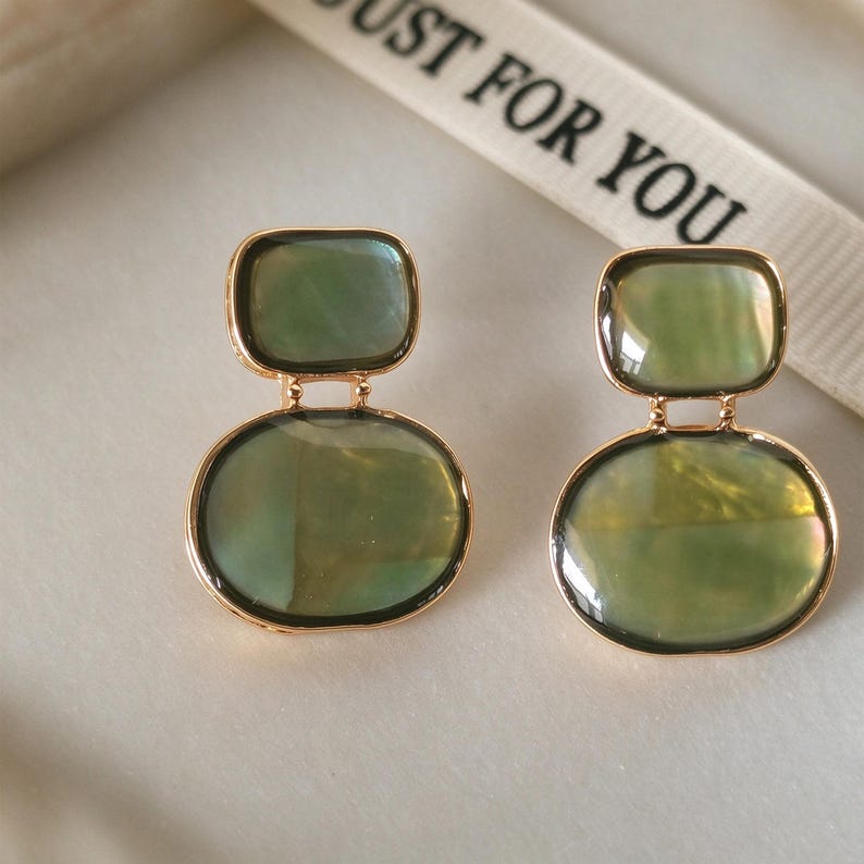 May include: A pair of gold-toned earrings featuring two stacked, smooth, olive-green iridescent stone elements. The top piece is rectangular with rounded corners, while the bottom piece is oval. The metal settings are thin and polished gold.