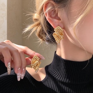 May include: A pair of gold-toned earrings featuring a textured, ribbon-like folded design. One earring is worn on the earlobe, while the other is held, showcasing a small cluster of clear, sparkling stones at the bottom edge of each piece.
