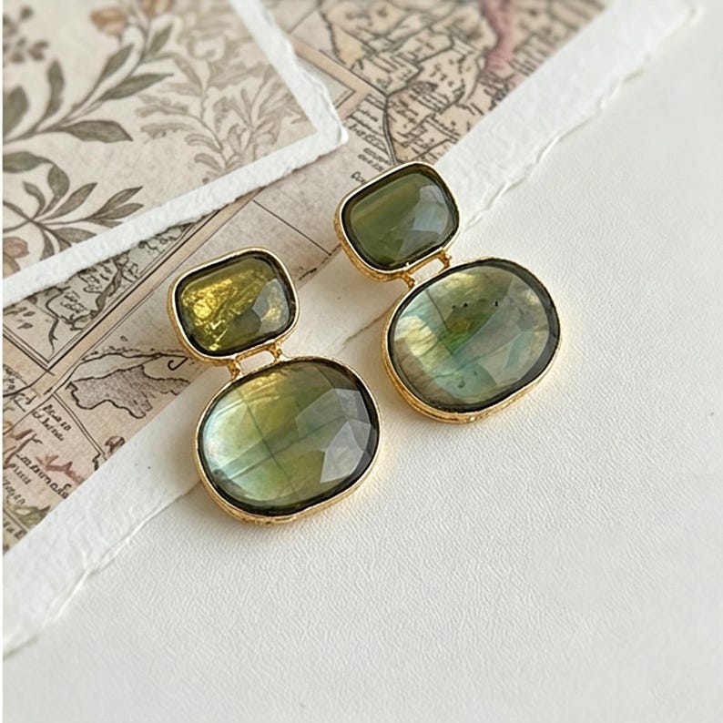 Close-up of handmade drop earrings with green gemstones and Gold-Tone details, placed on vintage-style paper and botanical background.