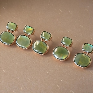 May include: Five pairs of earrings, each featuring two stacked, smooth, olive-green cabochon stones set in gold-toned metal. The top stone is a smaller rectangle, while the bottom stone is a larger oval, both with a subtle iridescent sheen.