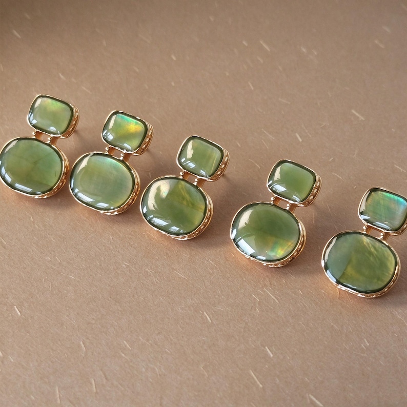 May include: Five pairs of earrings, each featuring two stacked, smooth, olive-green cabochon stones set in gold-toned metal. The top stone is a smaller rectangle, while the bottom stone is a larger oval, both with a subtle iridescent sheen.