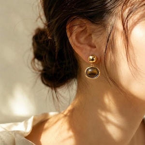May include: A gold-toned earring featuring a small polished sphere at the top connected to a larger, oval-shaped pendant. The pendant contains a smooth, brown, and tan striped gemstone with a chatoyant luster, set within a polished gold frame.