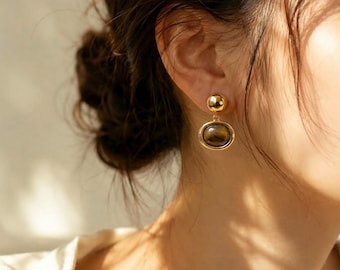May include: A gold-toned earring featuring a small polished sphere at the top connected to a larger, oval-shaped pendant. The pendant contains a smooth, brown, and tan striped gemstone with a chatoyant luster, set within a polished gold frame.
