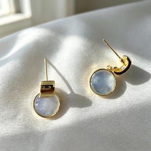 May include: A pair of gold-toned stud earrings featuring a circular, iridescent white shell inlay. Each earring has a polished gold metal setting with a post back, showcasing a smooth, reflective surface and a clean, minimalist circular design.
