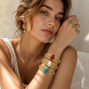 Puede incluir: Tres brazaletes de metal dorado con intrincados patrones de filigrana y cabujones ovalados centrales. El brazalete superior tiene una piedra roja, el central una cuenta blanca tipo perla y el inferior una piedra de color azul turquesa.