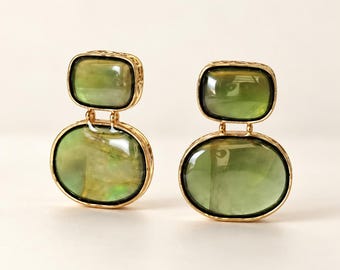 May include: A pair of gold-toned drop earrings featuring two stacked, smooth, translucent olive-green stones. The upper stone is rectangular with rounded corners, while the lower stone is oval. Each stone is encased in a textured gold metal frame.