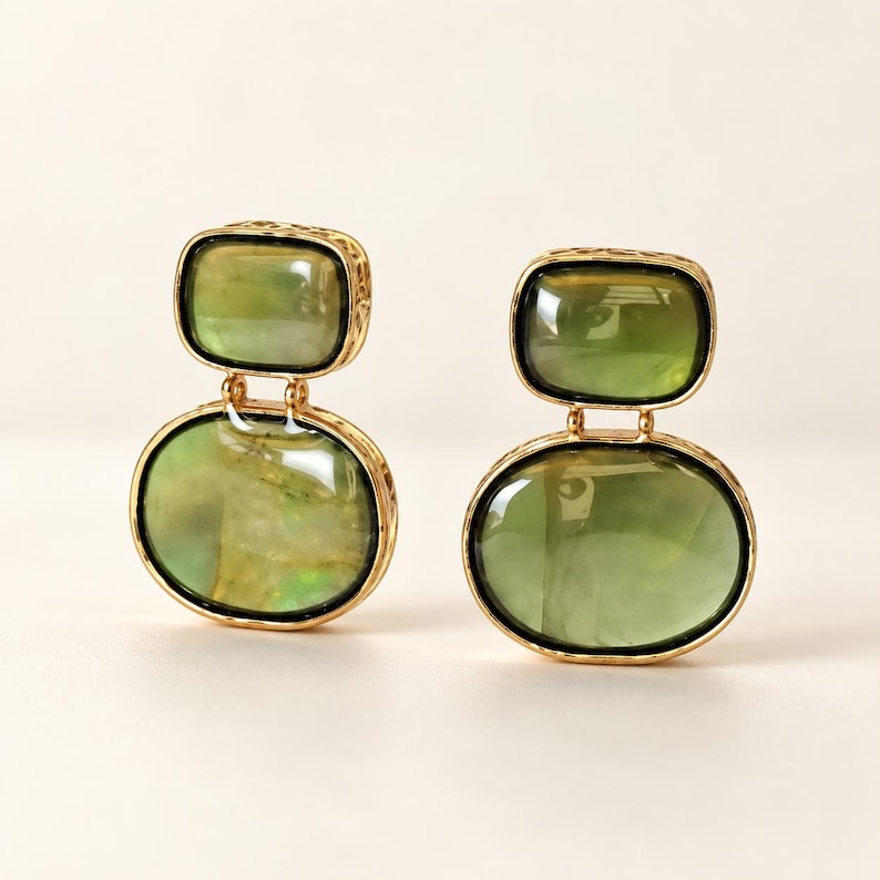 May include: A pair of gold-toned drop earrings featuring two stacked, smooth, translucent olive-green stones. The upper stone is rectangular with rounded corners, while the lower stone is oval. Each stone is encased in a textured gold metal frame.