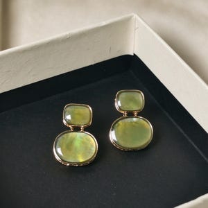 May include: A pair of gold-toned earrings featuring two stacked, smooth, translucent olive-green stones. The top stone is a smaller rectangle with rounded corners, while the bottom stone is a larger, rounded oval shape, both set in metal.