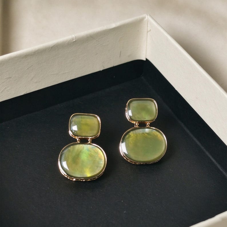 May include: A pair of gold-toned earrings featuring two stacked, smooth, translucent olive-green stones. The top stone is a smaller rectangle with rounded corners, while the bottom stone is a larger, rounded oval shape, both set in metal.