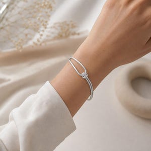 Puede incluir: Una pulsera de tono plateado con un diseño de cadena de doble hebra que se conecta a un solo bucle ovalado alargado. La pulsera se asegura con un mecanismo de cierre texturizado en forma de nudo que une las dos hebras en la muñeca.
