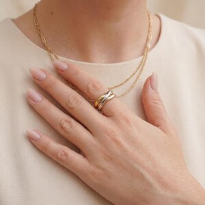 May include: A multi-tone interlocking ring featuring three smooth bands in yellow, rose, and white gold tones. The ring is worn on a finger, accompanied by a delicate layered gold chain necklace with a textured link design.
