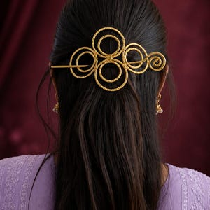 Könnte beinhalten: Ein goldfarbenes Haaraccessoire aus Metall mit einer Reihe von miteinander verbundenen Kreis- und Spiralformen mit strukturierter Oberfläche. Das dekorative Stück wird mit einer dünnen, geraden Metallnadel horizontal im Haar befestigt.