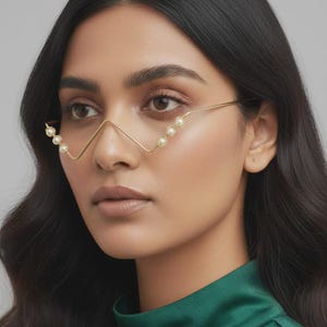 Könnte beinhalten: Goldfarbene Drahtbrillengestelle mit Kunstperlen. Die Brille hat ein einzigartiges, eckiges Design mit Perlenakzenten an den oberen und unteren Kanten. Das Model trägt ein grünes Satin-Top.