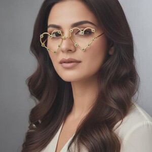 Könnte beinhalten: Goldfarbene Brille in Herzform, verziert mit kleinen grünen Edelsteinen. Die Brille wird von einer Person mit langen, welligen braunen Haaren und einem weißen Oberteil getragen. Der Hintergrund ist einfarbig grau.