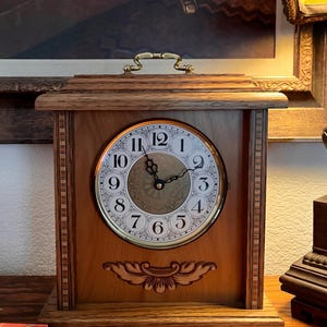 May include: A wooden mantel clock with a rectangular frame, a brass handle on top, and a circular face featuring black Arabic numerals from one to twelve. The center of the clock face has a gold-toned decorative pattern, and a carved wooden floral motif is centered below the dial.