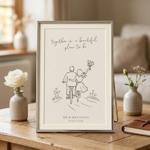 Peut inclure: Une impression encadrée présentant une illustration au trait de deux personnes faisant du vélo ensemble. Le texte indique "together is a beautiful place to be", "MR & MRS DAVIS" et "19 JUNE 2026". Le motif est en encre sombre sur fond crème.