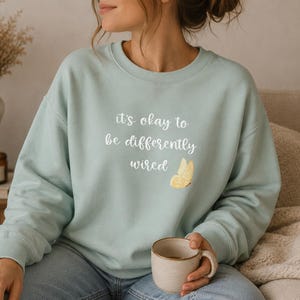 Könnte beinhalten: Hellblaues Sweatshirt mit dem Text "it's okay to be differently wired" und einer gelben Schmetterlingsgrafik. Das Sweatshirt ist aus weichem Material und für den Alltag geeignet.