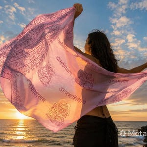 Può includere: Una sciarpa rosa chiaro con motivi e testo intricati è tenuta contro un tramonto sull'oceano. La sciarpa ha una qualità fluida e ariosa, con testo ed elementi decorativi visibili.