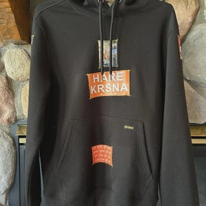 May include: Black hoodie with a drawstring hood and a kangaroo pocket. The front features the text "HARE KRSNA" in orange, with additional text and imagery. The hoodie is hanging on a metal hanger.