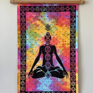 May include: A vibrant wall tapestry depicting a meditating silhouette with chakra symbols. The background is a tie-dye pattern in pink, yellow, blue, and green. It is framed with a black border and wooden hanging rods.