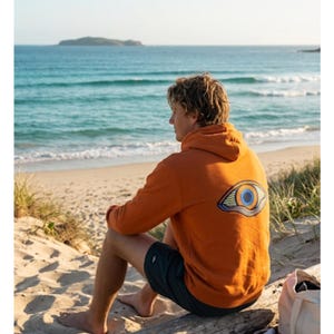 May include: An orange hoodie with a large eye design on the back. The hoodie is worn by a person sitting on a log at the beach. The person is wearing dark shorts. The background shows the ocean and a small island.