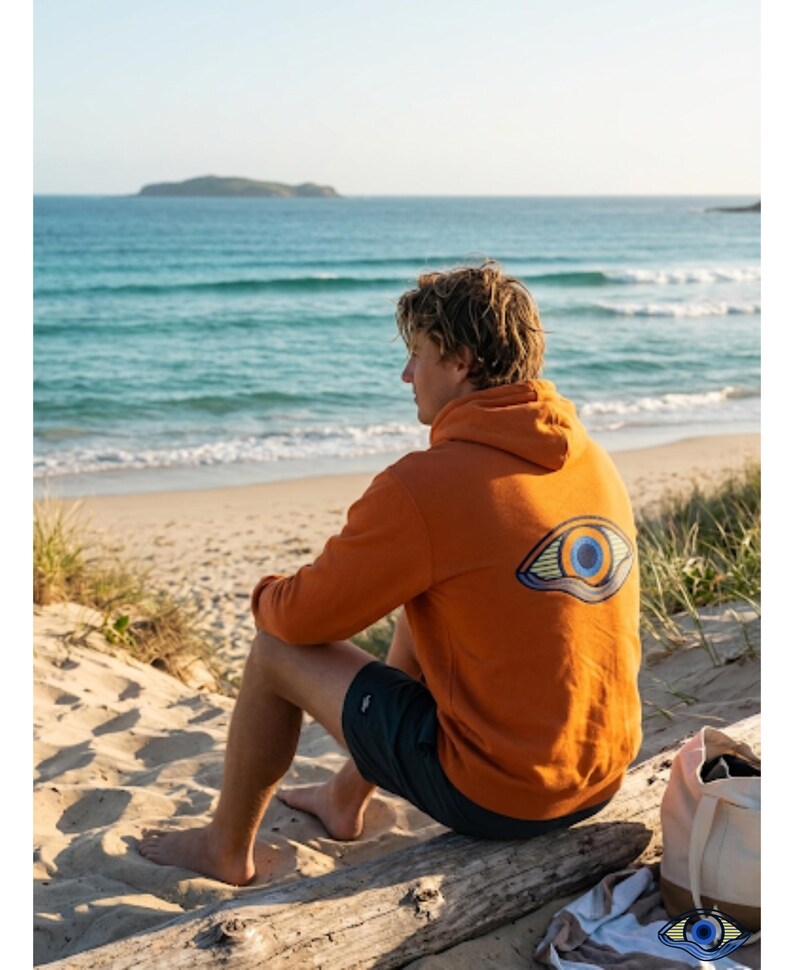 May include: An orange hoodie with a large eye design on the back. The hoodie is worn by a person sitting on a log at the beach. The person is wearing dark shorts. The background shows the ocean and a small island.