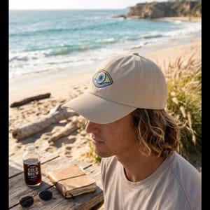 May include: Tan baseball cap with an embroidered eye design. The cap is worn by a person on a beach. The image also shows a bottle of cold brew coffee, a journal, and sunglasses on a wooden table near the ocean.