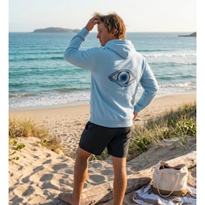 May include: A light blue hoodie with a large eye design on the back. The hoodie is worn with dark gray shorts. A beach bag and towel are on the sand. The person is standing on a sandy beach, looking out at the ocean.